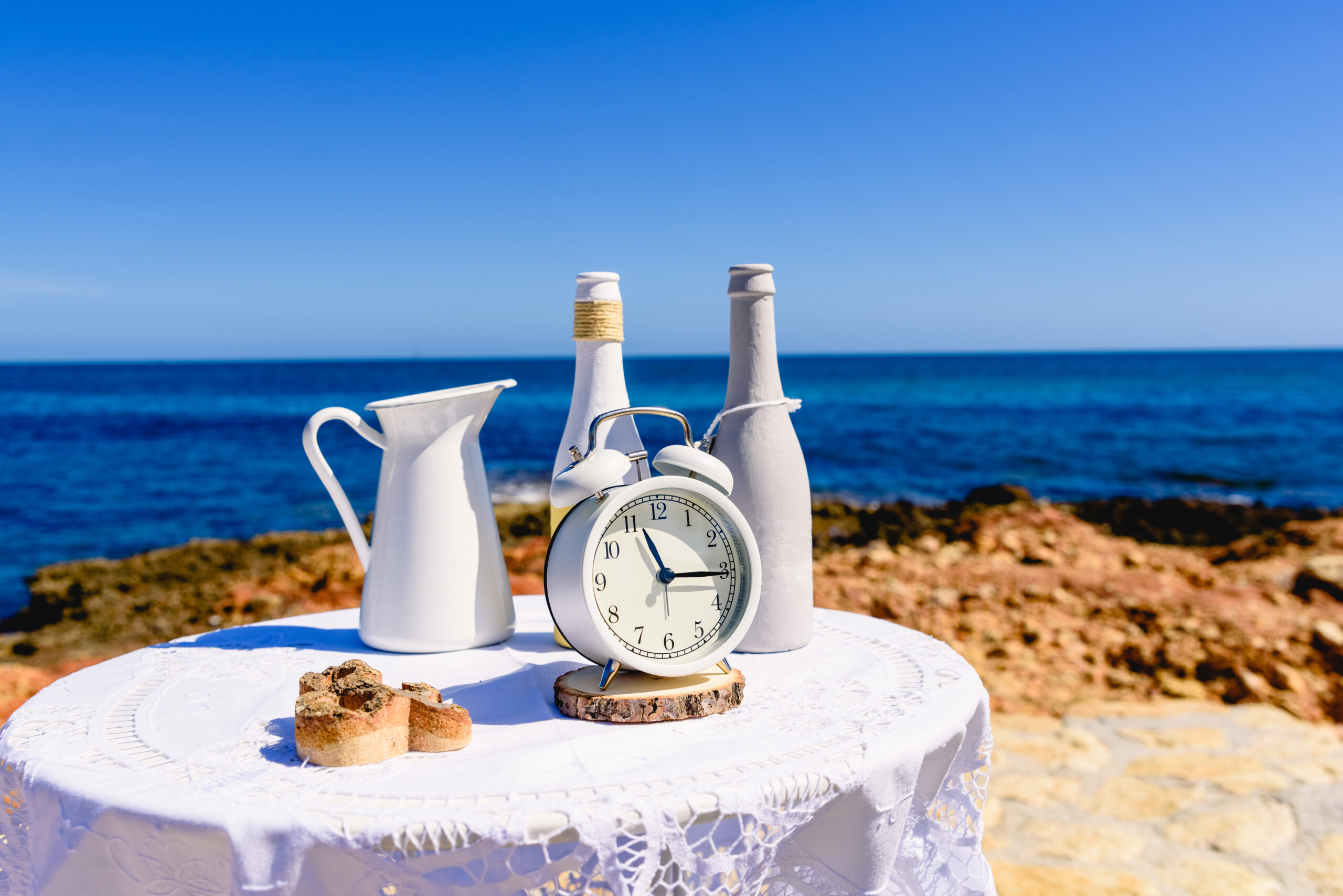 White table with antique clock placed on the rocks of the beach, with sea background and intense blue sky.